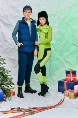 Sıcak giyimli şık ırklar arası çift hediyeler, kayaklar ve karlı stüdyoda noel ağacı yanında.