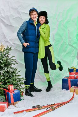 Kış kıyafetli ırklar arası bir çift Noel ağacının yanında gülümsüyor ve stüdyoda karda hediyeler sunuyor.