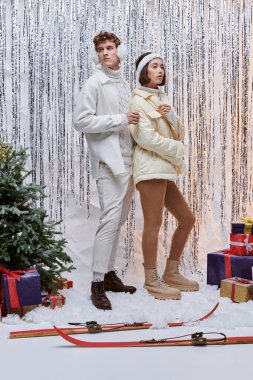 Kış kıyafetli ırklar arası bir çift stüdyoda, gümüş rengi süslemeler ve kar üzerinde noel hediyeleri.
