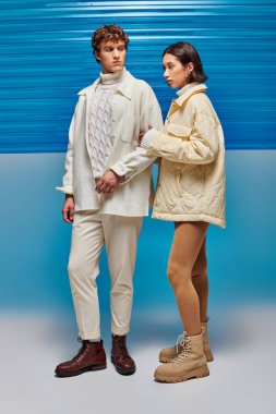 trendy interracial couple in warm outerwear posing on blue backdrop with plastic sheet, winter style clipart