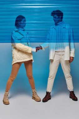 full length of stylish interracial couple holding hands behind blue plastic in studio, winter outfit clipart