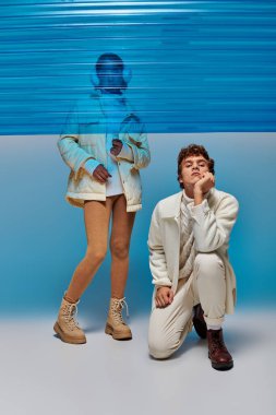 Sıcak elbiseli bir adam, mavi plastik panelin arkasındaki Asyalı kadının yanında, kış tarzında kameraya bakıyor.