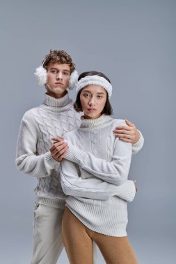 man in winter earmuffs embracing trendy asian woman in sweater on grey, fashion lookbook concept clipart