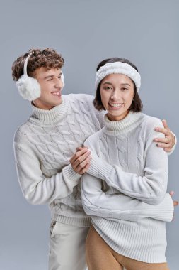 Sıcak kulaklıklı neşeli bir adam, gri, mevsimsel modaya uygun kışlık kazaklı Asyalı bir kadını kucaklıyor.