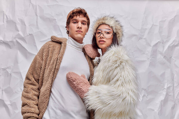 стильный мужчина и азиатская женщина в искусственной меховой куртке и рукавицах, смотрящих в камеру на белом бумажном фоне