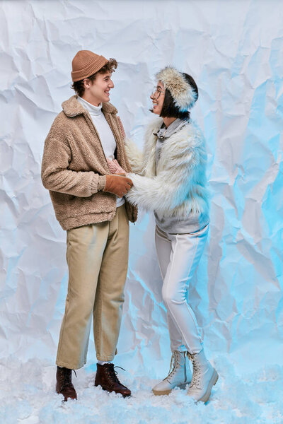 happy interracial couple in trendy winter clothes looking at each other on snow and white backdrop
