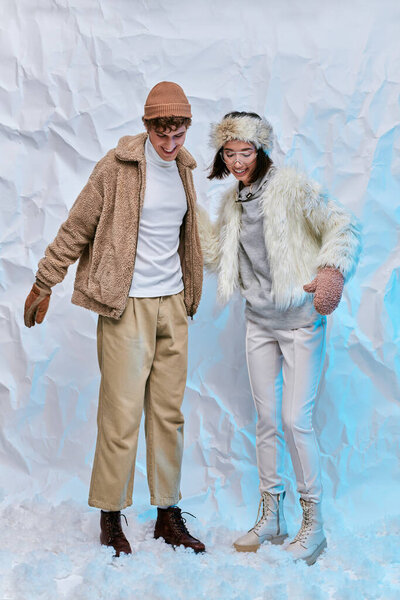 joyful interracial couple in trendy winter clothes standing on snow on white textured backdrop