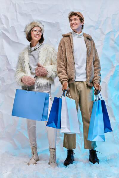 winter fashion campaign, happy interracial couple with blue shopping bags on white snow in studio