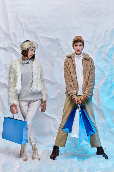 fashionable interracial couple with blue shopping bags standing on snow in studio, winter style