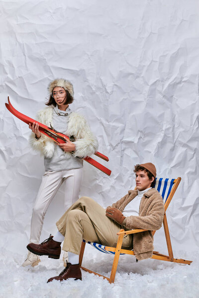 зимняя модная кампания, азиатка с лыжами рядом с модным мужчиной в шезлонге в студии на белом снегу