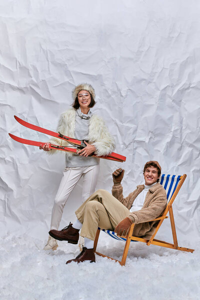 cheerful asian woman with skis near trendy man in deck chair on snow in studio, winter leisure