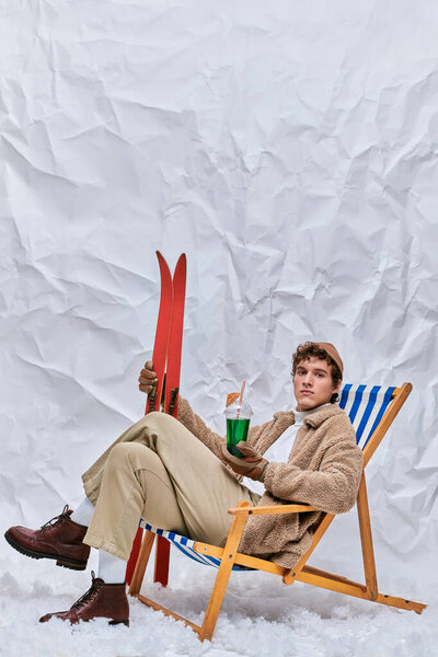 модный человек в теплой одежде сидит в шезлонге с горячим коктейлем и напитком в снежной студии