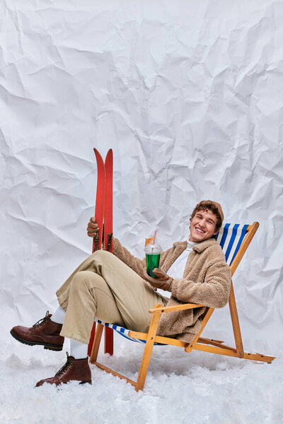 весёлый мужчина в зимнем наряде сидит в шезлонге с напитком и лыжами в снежной студии