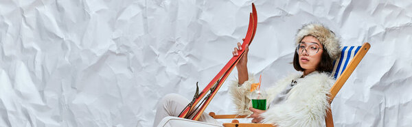 asian woman in winter attire in deck chair with skis and cocktail on white textured backdrop, banner