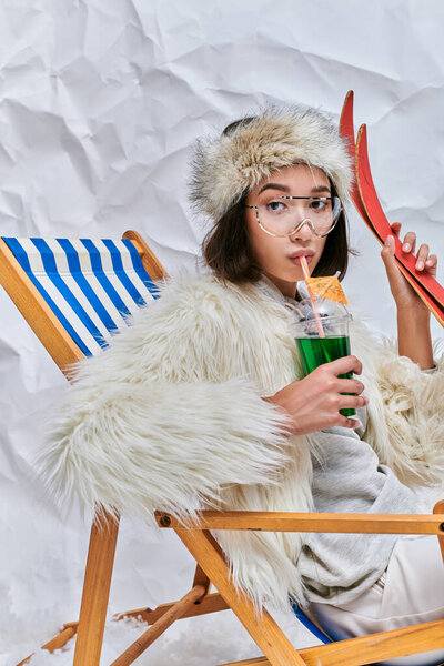 asian woman with skis drinking hot teddy cocktail in deck chair on white textured backdrop