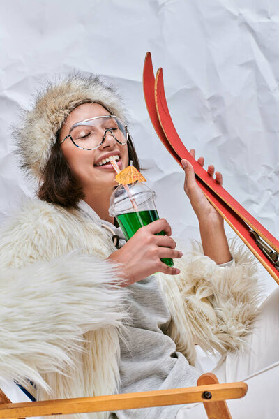 весёлая азиатская модель с лыжами, пьющими коктейль "apres-ski" в шезлонге на белом текстурированном фоне