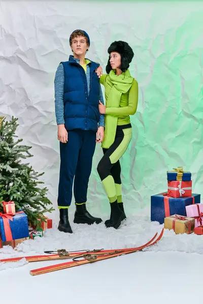 Stock image modern interracial couple in winter wear near presents, skis and christmas tree on snow in studio