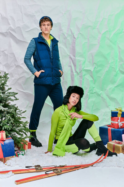 asian woman sitting on snow in studio near trendy man, skis and presents with christmas tree