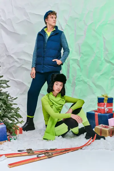 Stock image asian model sitting on snow in studio near stylish man, skis and gift boxes with christmas tree