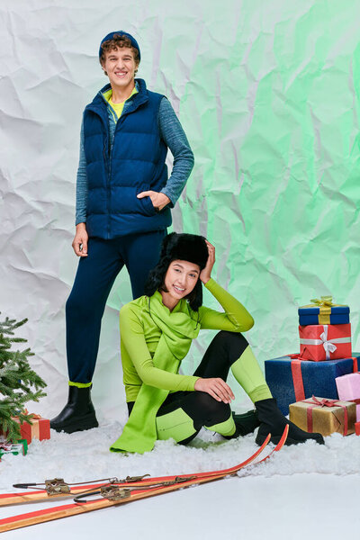 vibrant interracial couple posing on snow in studio near christmas tree, gift boxes and skis