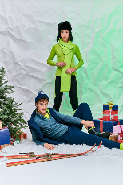 asian woman looking at camera near trendy man on snow near presents and christmas tree in studio