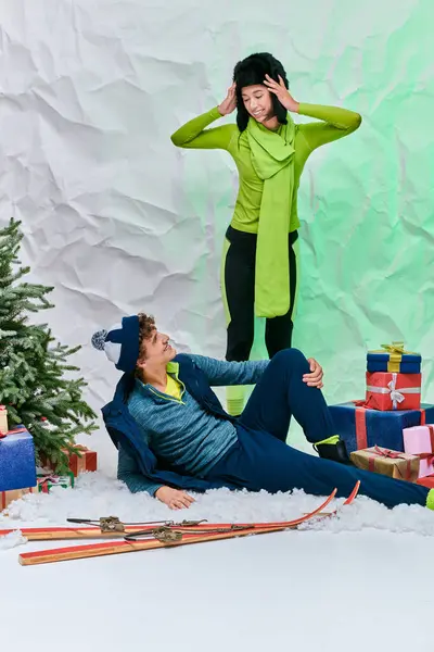 在工作室里 一对跨种族夫妇在雪地上的礼物 滑雪板和圣诞树旁互相微笑 — 图库照片