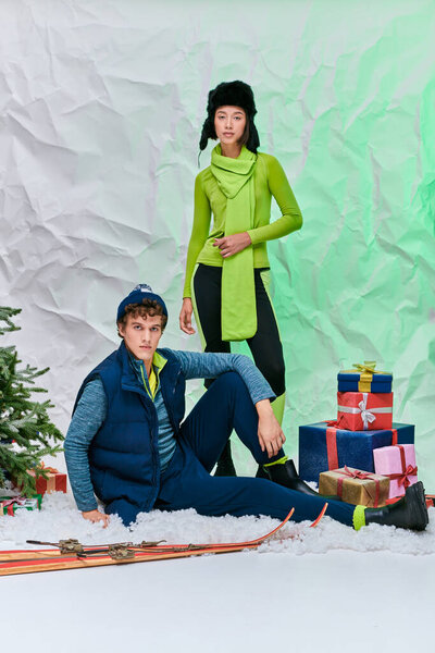 fashionable multiethnic couple looking at camera next to presents and christmas tree in snowy studio