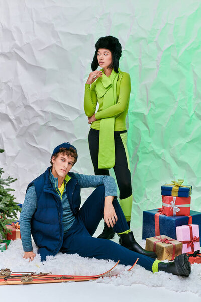 festive season, interracial couple posing on snow in studio near christmas tree and gift boxes