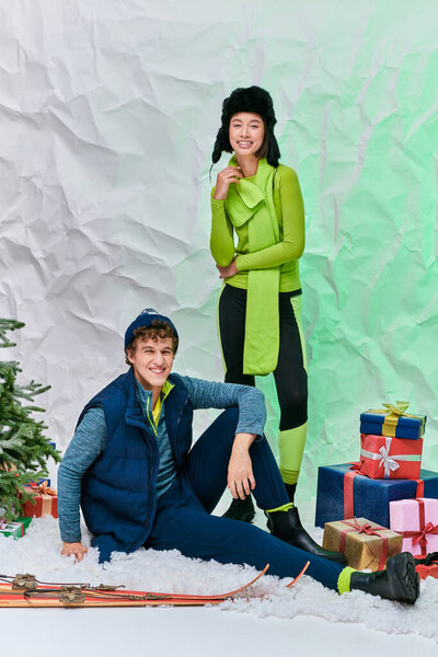 interracial couple in winter attire smiling near Christmas tree, skis and presents in snowy studio