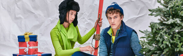 asian woman with skis looking at trendy man near christmas tree and presents in white studio, banner