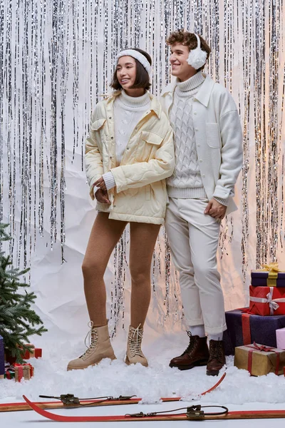 stock image multiethnic couple in winter clothes near present and christmas tree on shiny tinsel backdrop