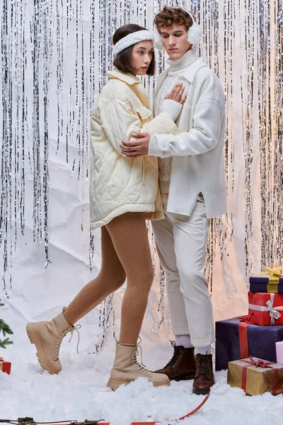 multiethnic couple in trendy clothes standing near Christmas presents on shiny tinsel backdrop