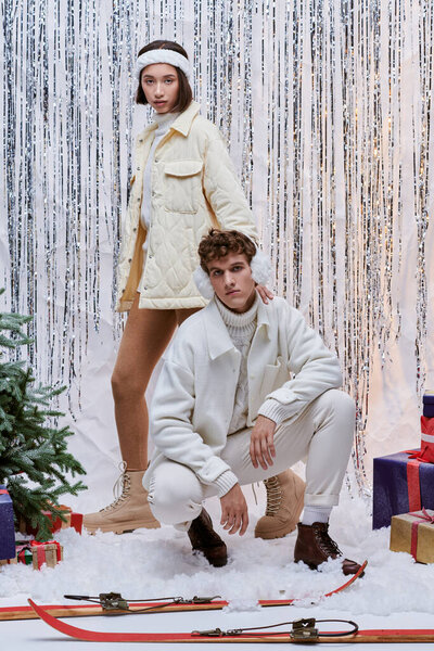 interracial models in winter clothes posing near christmas tree, presents and shiny decor in studio