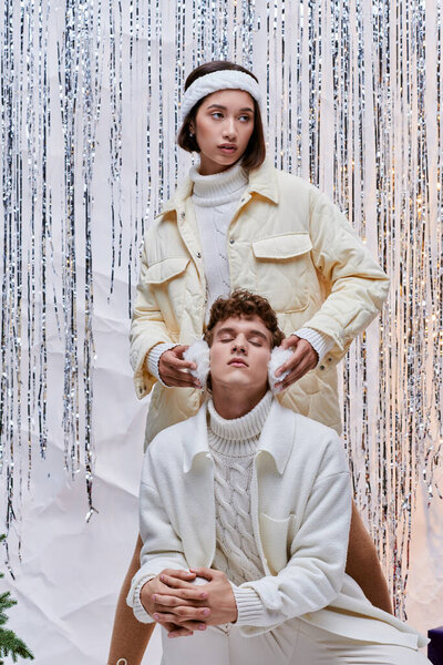 Азиатская женщина трогает теплые наушники модного мужчины, позирующего с закрытыми глазами рядом с серебряной мишурой