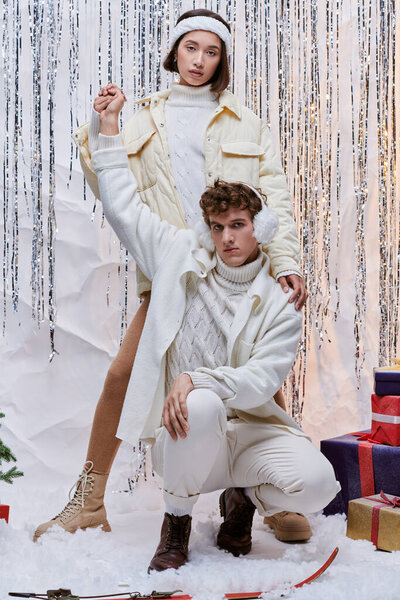 молодая азиатка держит за руку модного мужчину, позируя на снегу в студии рядом с серебряным декором