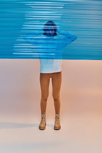 full length of young asian woman in white sweater and leather boots behind blue plastic