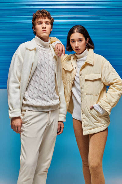 multiethnic couple in warm jackets and leather boots looking at camera on blue plastic backdrop