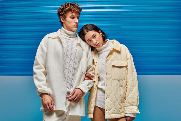 young asian woman leaning on shoulder of trendy man on blue plastic backdrop, winter style