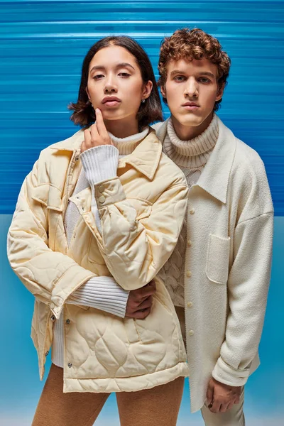 stock image winter fashion campaign, interracial couple jackets looking at camera on blue plastic backdrop