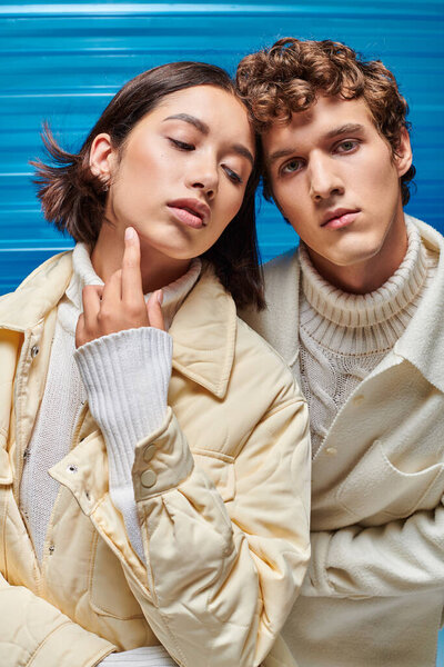 young man looking at camera near charming asian woman in winter outfit on blue plastic backdrop