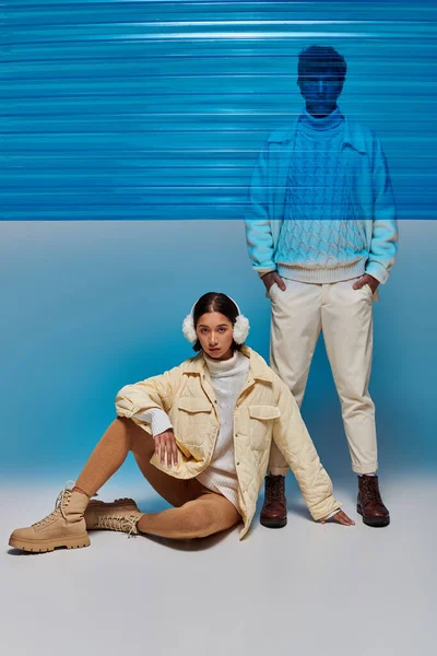 stock image asian woman in warm earmuffs sitting near man with hands in pockets and blue plastic sheet in studio