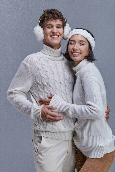 joyful multiethnic couple in knitted sweaters hugging under snowfall on grey backdrop, winter style