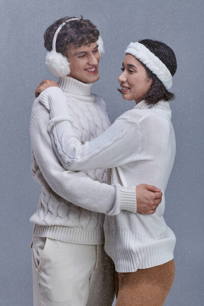 happy multiethnic couple in warm sweaters embracing under falling snowflakes on grey background