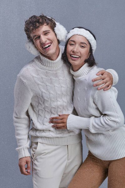 excited interracial couple in warm sweaters embracing and laughing under falling snow, happy winter