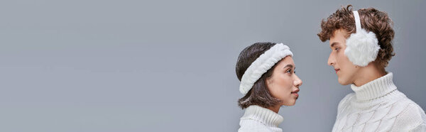 multiethnic couple in knitted sweaters and with snowy hair looking at each other on grey, banner