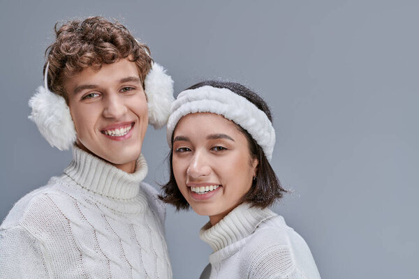 cheerful interracial couple in winter sweaters posing with snowy hair and looking at camera on grey