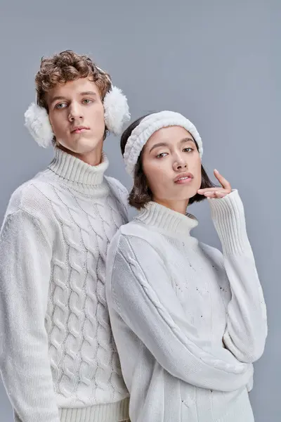 stock image young and stylish multiethnic couple in white cozy sweaters posing with snowy hair on grey backdrop