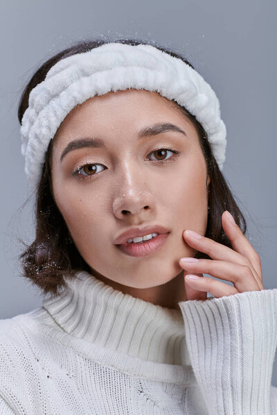 portrait of charming asian woman in warm headband touching face and looking at camera on grey