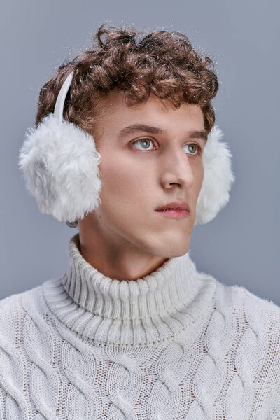 stylish man with snow on wavy hair, in earmuffs and white sweater looking away on grey, portrait