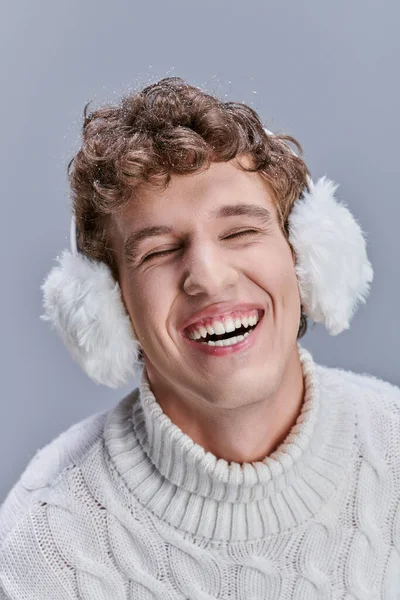 excited man in earmuffs and white sweater laughing with closed eyes on grey, happy stylish winter